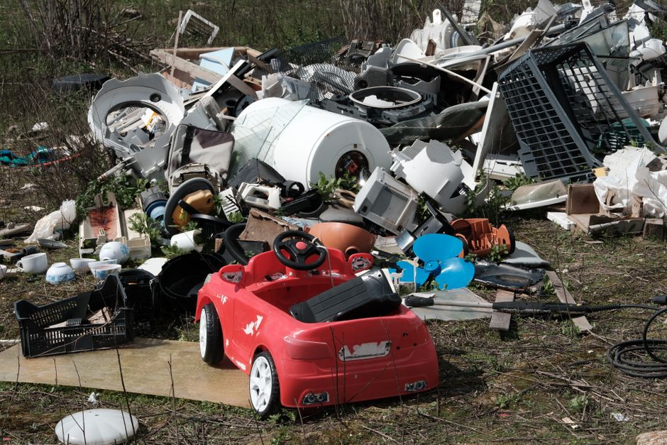 The image shows a large pile of mixed household waste and debris situated outdoors on a patch of ground with sparse grass and dirt. Prominently featured in the foreground is a small, red, plastic children's ride-on car with a slightly worn finish, visible scratches, and areas where the paint has chipped, revealing the white plastic underneath. Surrounding the toy are various discarded items including a black plastic crate, a blue inflated balloon, and a black extension cord coiled on the ground. In the background, the waste pile contains an assortment of broken appliances, such as a white washing machine drum, a microwave oven with visible internal components, and parts of kitchen fixtures, all haphazardly stacked and partially covered with rusted metal grills, plastic containers, and torn packaging. The scene is illuminated by natural daylight, with no visible signage or human activity, depicting an informal and unmanaged waste dumping site typical of private disposal or onsite clearance services like Waste Disposal Westminster, which manages rubbish collection outside of municipal systems.