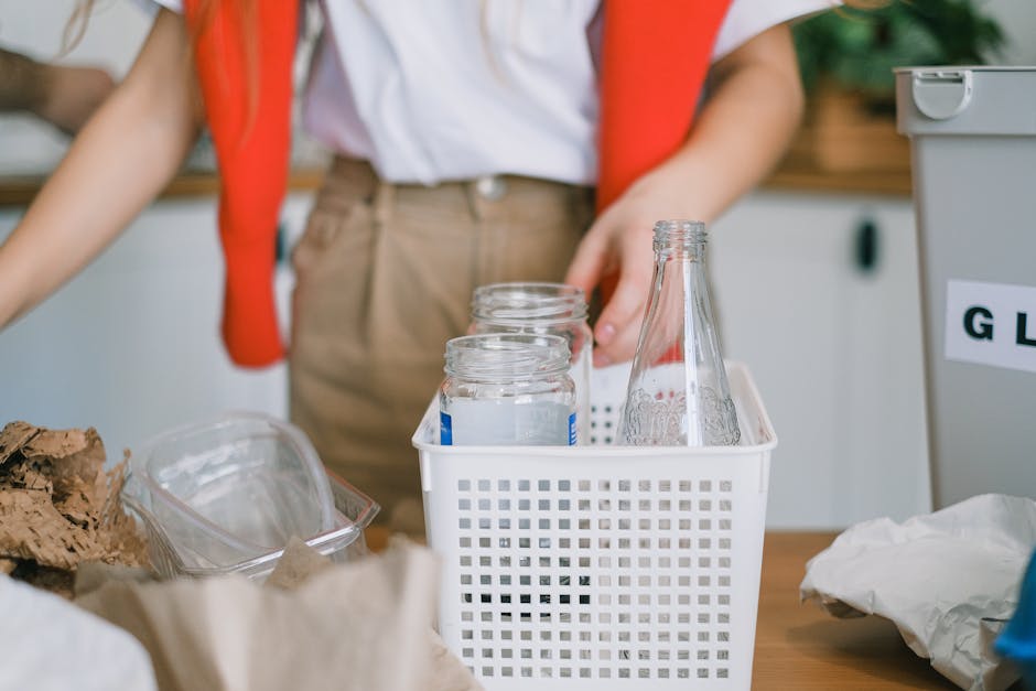 A young woman with long brown hair and light skin stands in a modern kitchen with a neutral grey wall and hanging utensils in the background. She is wearing a white T-shirt and has an orange sweater draped over her shoulders. She is holding a clear plastic water bottle in her right hand and appears to be unscrewing the cap or examining it closely. On the wooden countertop in front of her, there is a white plastic bin labeled 'PLASTIC' with a transparent lid, indicating it is used for sorting recyclables as part of a household waste management process. The scene suggests an on-site or private waste separation effort, supporting sustainable rubbish disposal practices. The lighting is soft and natural, highlighting the textures of the bottle’s smooth plastic surface and the fabric of her clothing, while the environment emphasizes organized, eco-friendly household rubbish collection within a domestic setting, aligning with the context of household rubbish pickup and waste disposal services. The overall atmosphere is clean, organized, and conducive to environmentally responsible waste handling.