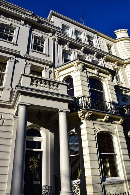 A detailed view of a multi-storey Victorian-style residential building with ornate architectural features, including white-painted stonework, decorative cornices, and black wrought iron balcony railings. The building's facade is primarily light cream with contrasting dark window frames and balcony supports. Large sash windows with arched tops are visible on the lower floors, some adorned with wooden blinds, while smaller rectangular windows are set into the upper levels. The entrance at ground level has a black front door with a decorative wreath hanging on it, flanked by two white columned porch supports. The building is situated on a paved street, with clear blue sky overhead, and appears to be part of an affluent urban area. This type of architecture may be associated with independent property ownership or private rentals, often requiring professional waste disposal services such as those offered by Waste Disposal Westminster, especially for maintenance or renovation waste during on-site clearance or refurbishments.