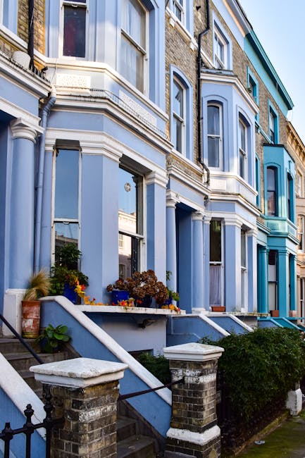 The image displays a row of Victorian-style terraced houses painted in various shades of blue, with decorative white trim and ornate architectural details. The front facades feature large sash windows, some with potted plants and flowers placed on small ledges beneath them. Concrete steps lead up to the entrance doors, which are situated behind brick pillars topped with concrete caps, and are accessed via short, narrow staircases with metal railings. The environment appears to be a residential street with well-maintained exteriors, and the sky is partially visible at the top of the image, indicating natural daylight. The scene exemplifies typical urban Victorian housing with a focus on decorative features and vibrant paintwork, with the presence of subtle greenery. This setting provides context for private property management or on-site clearance activities, which Waste Disposal Westminster in Westminster might facilitate as part of rubbish removal or estate clearance services related to similar residential areas.