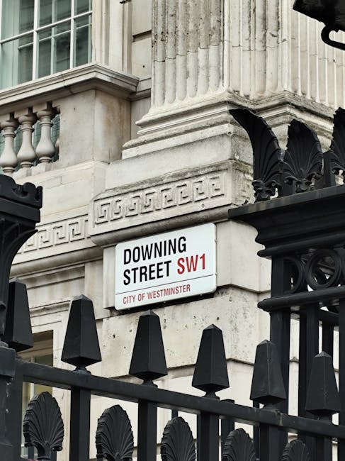 A white street name sign reading 'Downing Street SW1' with red and black text, mounted on a historic stone building featuring decorative column details and a classical architectural style. The sign is positioned above a black wrought iron fence with pointed finials and decorative elements, which partially obscures the lower part of the building façade. The background includes part of the stone façade and classical architectural features such as a pilaster and cornice, indicating a prominent government or official building in Westminster. The scene is illuminated by natural daylight, highlighting the textures and finishes of the stone and metal elements, typical of central London’s historic architecture. The context of the scene suggests an alternative or private approach to waste management at this location, aligning with the theme of rubbish removal and site clearance in a high-profile area managed by Waste Disposal Westminster.