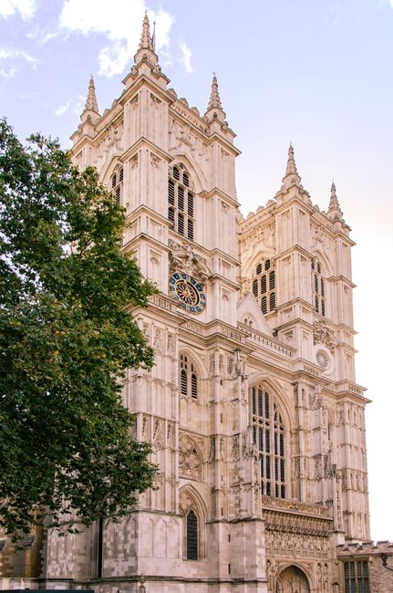 A detailed, upward-view of the historic Westminster Abbey building showing its Gothic architecture, with tall, pointed spires and ornate stone carvings. The facade features large, arched stained glass windows framed by intricate tracery and decorative stonework. The structure is constructed from light-colored stone that appears weathered but well-maintained. A prominent clock with a blue face and gold Roman numerals is situated near the center of the facade, just below the upper level of windows. The scene is set against a partly cloudy sky with patches of blue visible, and a leafy tree occupies the lower left corner, partially obscuring the base of the building. The photograph emphasizes the scale and detail of the abbey, with natural lighting highlighting the textures of the stone. Such a setting can be associated with historic landmarks often considered in the context of private or alternative disposal methods, such as specialist waste handling for heritage sites, aligning subtly with the theme of alternative waste management options for significant structures.