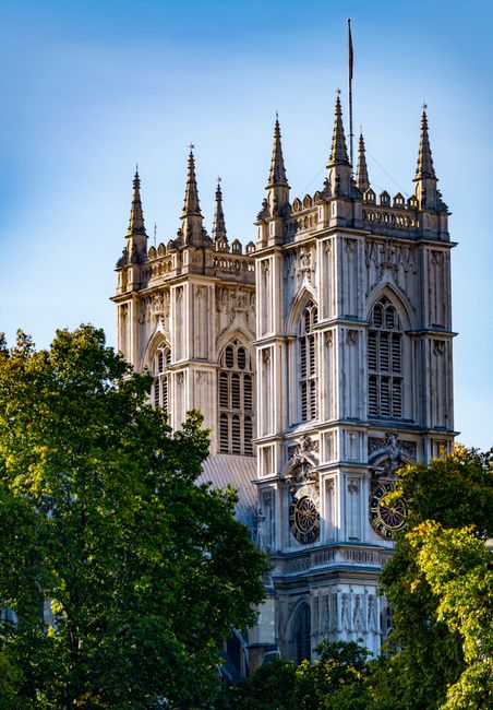This image shows the upper section of Westminster Abbey's Gothic-style twin towers built from light-colored stone, featuring ornate detailing, lancet windows, and decorative pinnacles. The towers are positioned prominently against a clear blue sky, with a flagpole flying a small flag at the top of the right tower. Surrounding the lower part of the image are lush green trees with dense foliage, partially obscuring the base of the towers. The stonework exhibits a textured surface with visible vertical and horizontal lines, emphasizing the architectural intricacies of the historic structure. The lighting in the scene suggests it is late afternoon or early evening, casting gentle shadows that highlight the depth and contours of the stone surfaces. Although the image centers on the abbey, the presence of greenery reflects the typical setting of the site amidst surrounding parkland or open spaces, often linked to private or non-municipal preservation efforts, aligning with the context of alternative waste handling and independent collection services like those offered by Waste Disposal Westminster.