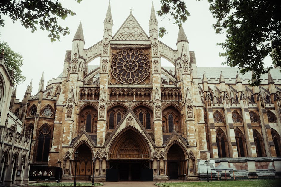 A detailed, upward-view of the historic Westminster Abbey building showing its Gothic architecture, with tall, pointed spires and ornate stone carvings. The facade features large, arched stained glass windows framed by intricate tracery and decorative stonework. The structure is constructed from light-colored stone that appears weathered but well-maintained. A prominent clock with a blue face and gold Roman numerals is situated near the center of the facade, just below the upper level of windows. The scene is set against a partly cloudy sky with patches of blue visible, and a leafy tree occupies the lower left corner, partially obscuring the base of the building. The photograph emphasizes the scale and detail of the abbey, with natural lighting highlighting the textures of the stone. Such a setting can be associated with historic landmarks often considered in the context of private or alternative disposal methods, such as specialist waste handling for heritage sites, aligning subtly with the theme of alternative waste management options for significant structures.